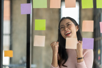 Confident businesswoman wearing eyeglasses showing joyful gesture on post-it note on glass wall in strategic business and online to achieve the goals set.
