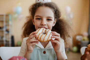 Front view portrait of cute little girl eating big donut during birthday party and looking at camera happily.