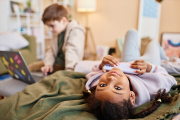 Wall Mural - Portrait of black girl looking at camera upside down while lying on bed with smartphone, copy space