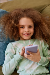 Wall Mural - Vertical top view portrait of cute girl looking at camera while lying on bed with smartphone