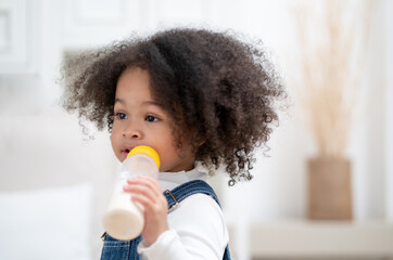 Cute curly hair biracial little girl sucking milk from bottle. Hungry kid holding milk bottle happy eating at home. Innocence childhood lifestyle.