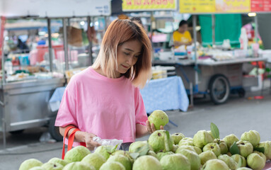 Asian woman standing at the local market and choosing buying fruit guava and holding bag plastick,organic food local market concept.