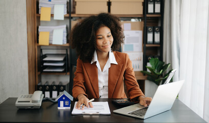 Young real estate agent worker working with laptop and tablet at table in office and small house beside it.