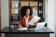 © Nuttapong punna - Confident beautiful African businesswoman typing laptop computer and digital tablet while holding coffee at office.