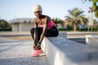 © Bojan - Young fitness afro-american woman with blond hair in sportswear tying shoe laces while sitting on concrete wall outdoors