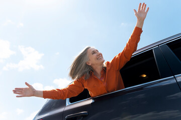 portrait of happy senior gray haired woman sitting inside new car, holding hands up, having fun enjo