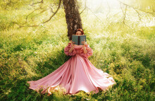 Fantasy Woman Sits Under Tree Holding Romantic Book In Hands Reading Novel. Pink Long Vintage Dress Fairy Princess Girl In Garden Summer Nature Green Grass Magic Sun Rays Light. Art Photo Real People