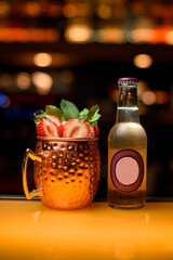 Wall Mural - Cocktail with ice, strawberries, mint in copper mug and bottle of ginger ale on bar counter