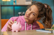 © Andrii Iemelianenko - Caucasian preteen school girl putting one dollar cash money into piggy bank. Young child enjoying her first saving at home workplace sitting at the table. Kids and financial literacy budget planning