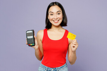 Young happy woman of Asian ethnicity she wear casual clothes red tank shirt hold wireless modern bank payment terminal to process acquire credit card isolated on plain pastel light purple background.