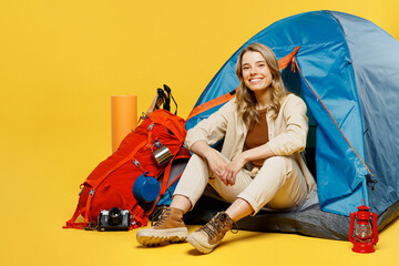 Full body smiling fun young woman sit near bag with stuff tent look camera isolated on plain yellow background. Tourist leads active lifestyle walk on spare time. Hiking trek rest travel trip concept.