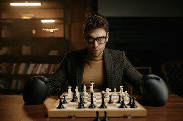 Angry young man wearing boxer gloves sitting at table with chessboard