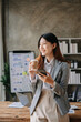 © Nuttapong punna - Confident beautiful Asian businesswoman typing laptop computer and digital tablet while holding coffee at office.