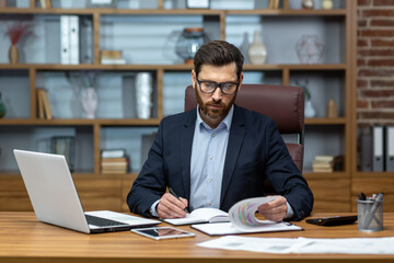 Serious and focused financier accountant on paper work inside office, mature man using calculator and laptop for calculating reports and summarizing accounts, businessman at work.