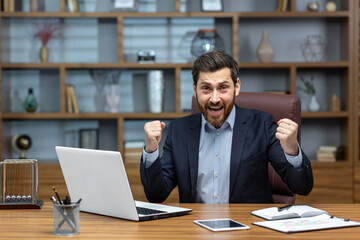 Portrait of mature senior businessman in office, man looking at camera and holding hands up super power and superhero gesture, successful investor using laptop inside building.