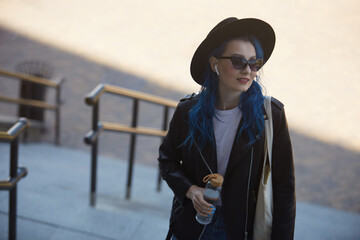 Wall Mural - Young adult female with blue hair walking on stairs in the city center with a reusable glass bottle of water and cottong shopper bag on shoulder. Sustainability and diversity concept