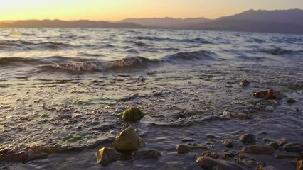 Sticker - Closeup of sea tide splashing on mossy stones with sunset orange sky