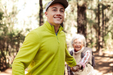 Handsome boy helps his grandmother to hike in the forest. Smiling multigeneration family couple enjoying mountain and nature together. The new generation helps the old one