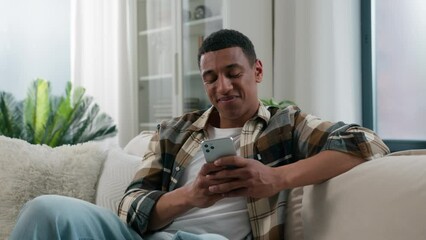 African American guy ethnic happy smiling man relaxing on comfortable couch sofa at home using mobile application scrolling phone app chatting social network online web surfing internet smartphone net