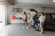 © BASILICOSTUDIO STOCK - Son plugs EV charger from charging station to electric vehicle in private home while mother takes groceries from the car on daytime. Sustainable Alternative Lifestyle. Horizontal copy-space.