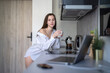 © Mindaugas Dulinskas - Beautiful Young Girl is Standing in the Kitchen and Drinking Coffee. Morning Routine. Wearing White shirts. Blurry Laptop and Mobile Phone on the Desk.