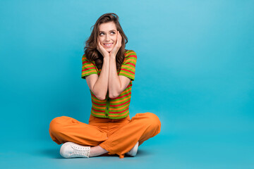 Poster - Full size photo of minded woman dressed orange pants colorful cardigan look empty space arms cheekbones isolated on blue color background