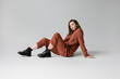 full length of trendy model with brunette and wavy hair sitting in trendy and oversize suit with terracotta blazer, pants and black boots, looking at camera on grey background, stylish young woman