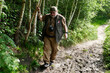 Active senior man of 70 with camera and walking stick tries to keep his shoes out of the mud and grabs a tree trunk