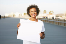 Medium Shot Portrait Photography Of A Grinning Woman In Her 50s Holding An Empty White Blank Sign Poster Wearing A Casual T-shirt Against A Beach Boardwalk Or Promenade Background