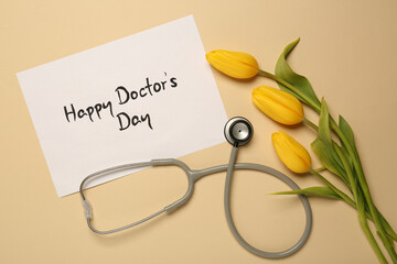 Wall Mural - Card with phrase Happy Doctor's Day, stethoscope and yellow tulips on beige background, flat lay