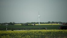 Cars Drive Along Highway As Wind Turbine Spins In Background In Yellow Farm Field