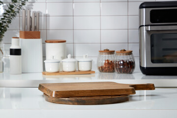 Stacked of wooden cutting board form an empty podium for display product. In the background is the counter with spice boxes, knife tray and microwave decorated on white tile background