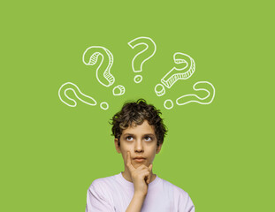 An 11-year-old boy, touching his chin with a dubious expression and looking up as if thinking, isolated on a green background with graphic question marks around him