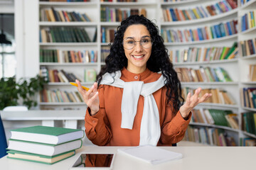 Webcam view online video call, young female student talking to friends remotely, Hispanic woman inside academic library at desk studying and preparing for exams.