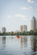 back view of interracial sportive couple in life vests sailing in kayak along riverside with green trees and contemporary buildings during recreation activity on summer weekend