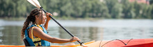 Side View Of Active And Happy African American Woman In Life Vest Holding Paddle And Sailing In Kayak While Spending Summer Weekend On Scenic River, Banner