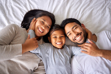 Wall Mural - Portrait, love and family in bed from above happy, smile and bonding in their home together. Face, girl child and top view of parents with their daughter in a bedroom, relax and embracing indoors