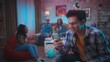 © kinomaster - A group of young people have fun at a house party, birthday party, drink drinks from paper cups. A curly haired guy in a festive hat sits with a phone and smiles joyfully.