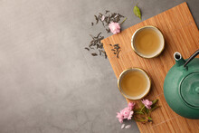 Traditional Ceremony. Cups Of Brewed Tea, Teapot And Sakura Flowers On Grey Table, Flat Lay With Space For Text