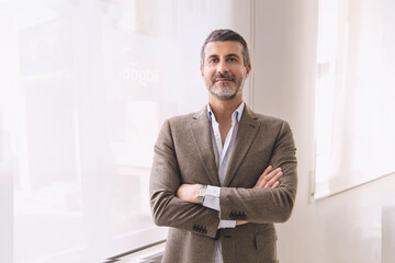 Confident Businessman in Bright Office Setting - Portrait of mature businessman with crossed arms, standing in white office illuminated by large window, in his forties, looking at camera