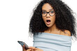 Close up portrait of stupefied young woman with Afro hairstyle, keeps mouth widely opened, holds smartphone, receives shocking message, wears blue sweater, shows bare shoulders, poses indoor