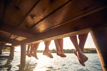 Wall Mural - Focus on legs of young people. Four young friends sitting on jetty by water. Rear view of legs hanging down. Travel, holiday, togetherness, fun, lifestyle concept.