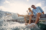 Fototapeta Na sufit - A young couple splashing their legs in the water while sitting at the dock on the seaside. Love, relationship, holiday, sea