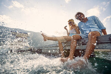 A Young Couple Splashing Their Legs In The Water While Sitting At The Dock On The Seaside. Love, Relationship, Holiday, Sea