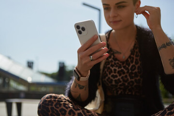 Wall Mural - Young woman in leopard clothes listening to music in headphones and browsing an application on a modern smart phone. Tattooed female person sitting on a bench and using a cellphone