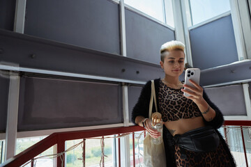 Wall Mural - Cool young diverse woman with short hair standing on stairs indoors and using a smart phone. Beautiful white female person holding a glass bottle of water in hand and wearing a tote bag on shoulder