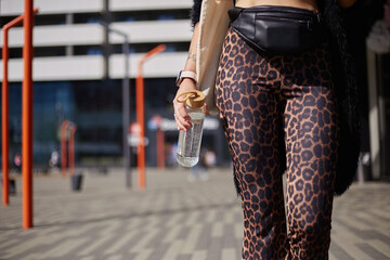 Wall Mural - Tattooed young woman in leopard clothes walking outdoor with a glass water bottle in hand. Unrecognizable female person leading a sustainable lifestyle