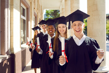 Wall Mural - College graduates standing behind each other and smiling. Group of happy international students in black mortar board caps and bachelor gowns holding diplomas. College or university education