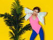 young fashion girl in iridescent jacket, pink body and blue leggings posing near green palm isolated on yellow background