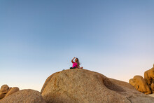 Woman, Tourist Seen In Joshua Tree National Park At Sunset Sitting On Top Of A Rock Boulder In Bright Pink Shirt Black Shorts. Blue, Orange Gradient Sky For Travel, Tourism, Influencer Shot.